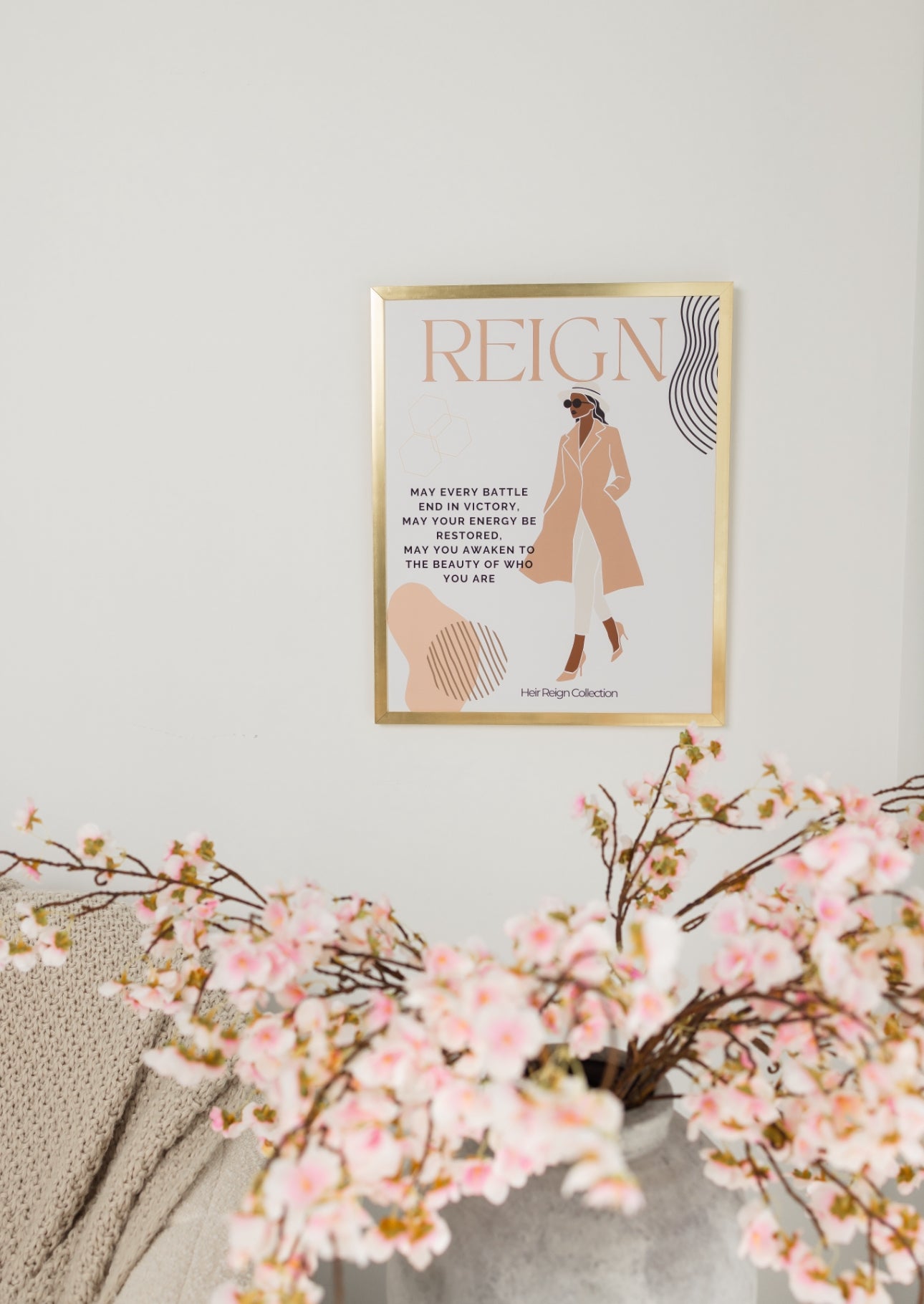 Beautiful wall art illustration of a black woman. She is wearing a pink trench coat, white  pants and high heel shoes. The picture also has an inspirational positive affirmation next to the woman. The picture is in a  gold frame with beautiful home decor. 