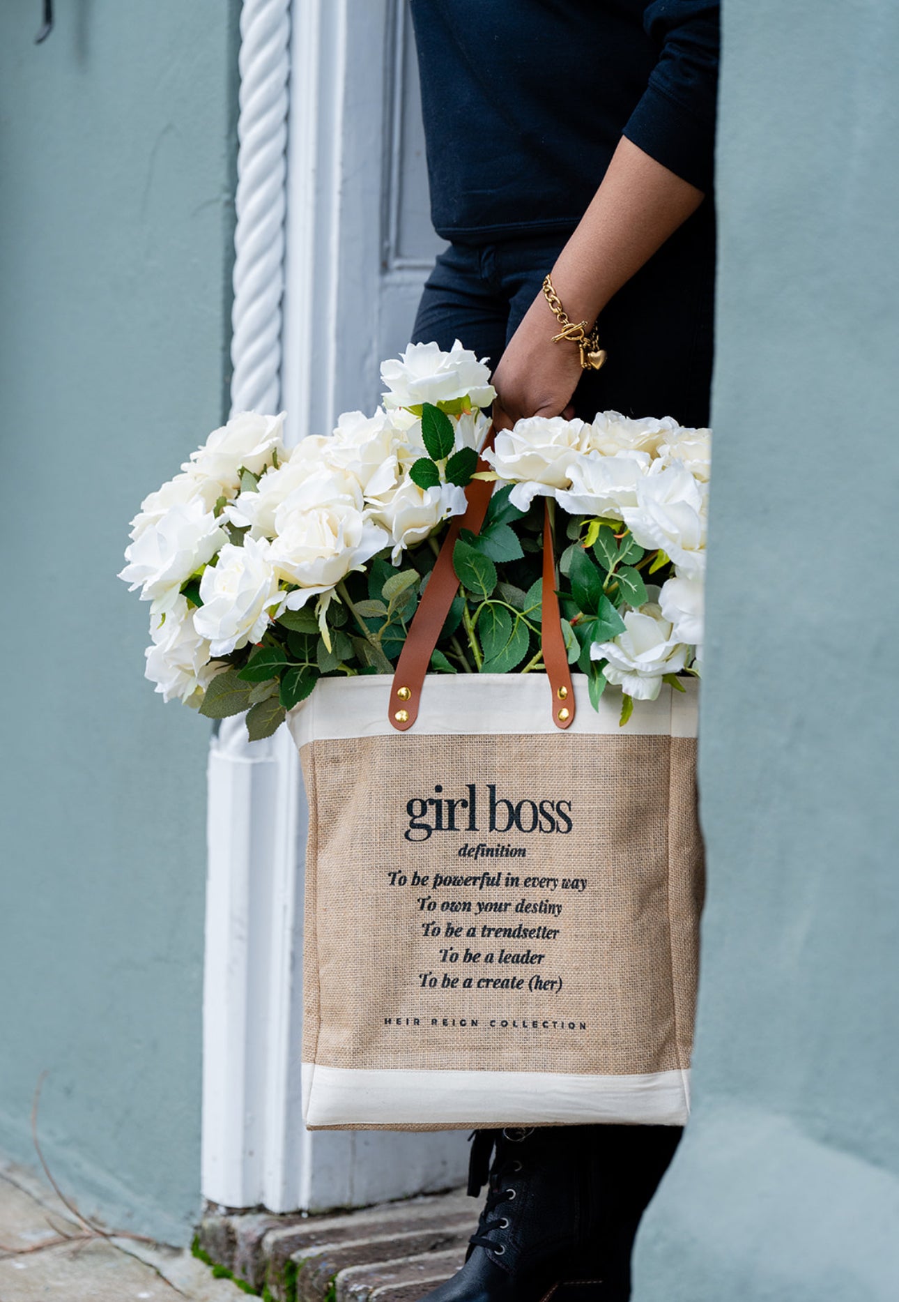 This is a picture of a burlap tote bag with a positive affirmation quote on the front. There is a woman holding the tote bag that has bright white summer flowers inside of the bag.