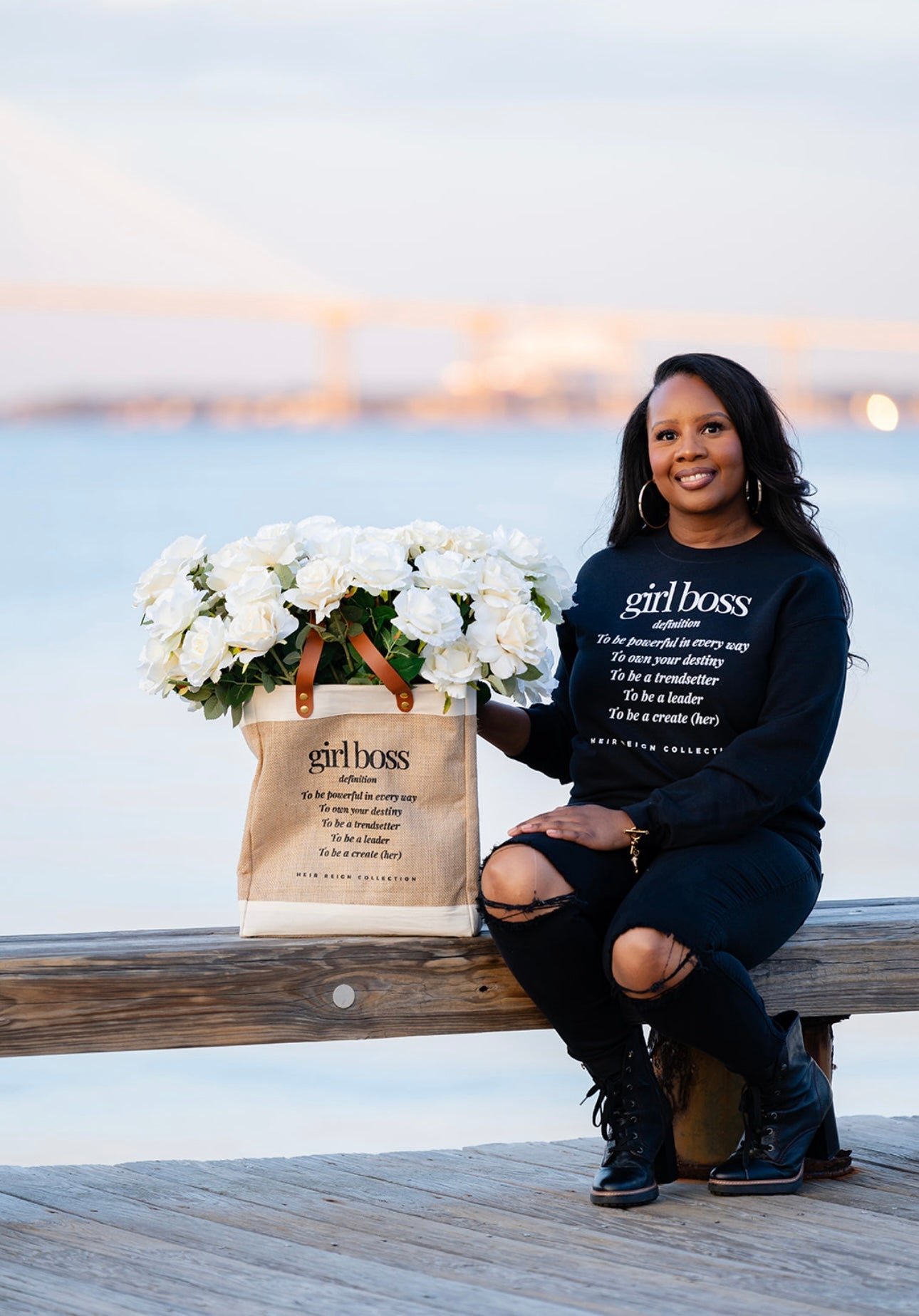 This is a picture of a burlap tote bag with a positive affirmation quote on the front. There is a woman holding the tote bag that has bright white summer flowers inside of the bag.